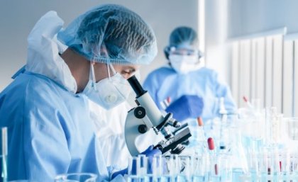 A scientist in cap, gown and mask stands in profile looking down through a microscope as a second scientist looks on in the background. They are in a lab.